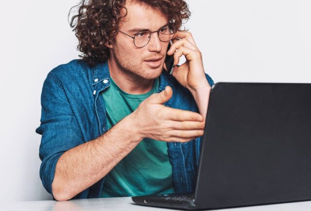 Mężczyzna przed komputerem wskazuje na monitor z wyraźnym niezadowoleniem. W lewej ręce trzyma przy uchu telefon.