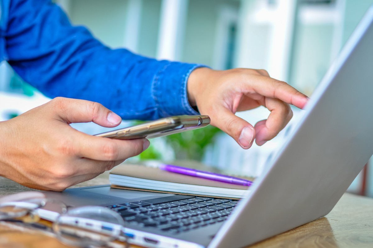 Zbliżenie na człowieka, który obsługuje laptopa oraz telefon.