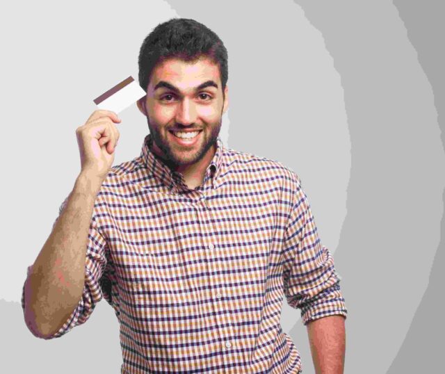Uśmiechnięty mężczyzna w koszuli w kolorową kratę trzymający kartę płatniczą lub kredytową w dłoni. Osoba stoi na jasnym tle i prezentuje kartę w geście zadowolenia.