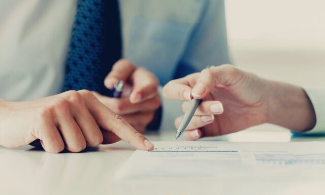 Zbliżenie na dłonie dwóch osób. Jedna wskazuje na fragment dokumentu, w momencie gdy druga popdisuje dokument.