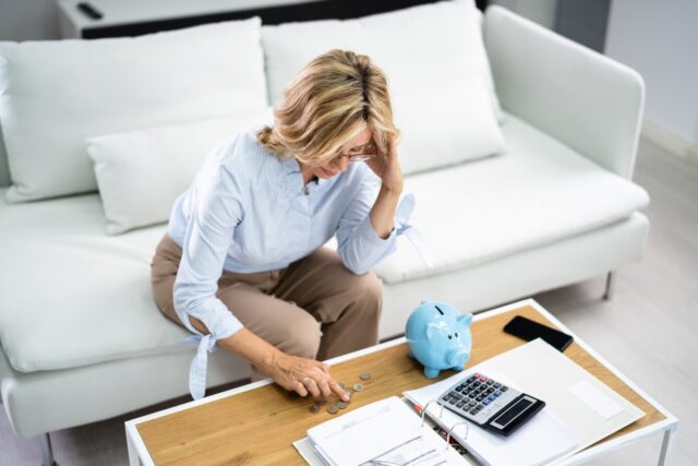 Kobieta siedząca na sofie liczy niewielką ilość pieniędzy na stole. Jej mina wskazuje na problemy z finansami.