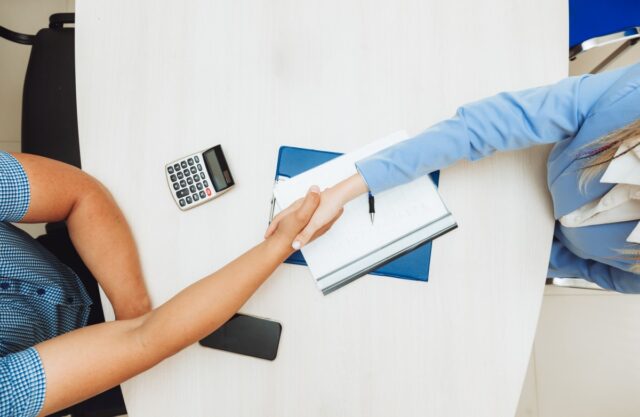 Dwie osoby podają sobie ręce nad dokumentami i kalkulatorem. Może to wskazywać na podanie sobie ręki na przypieczętowanie zgody biznesowej.