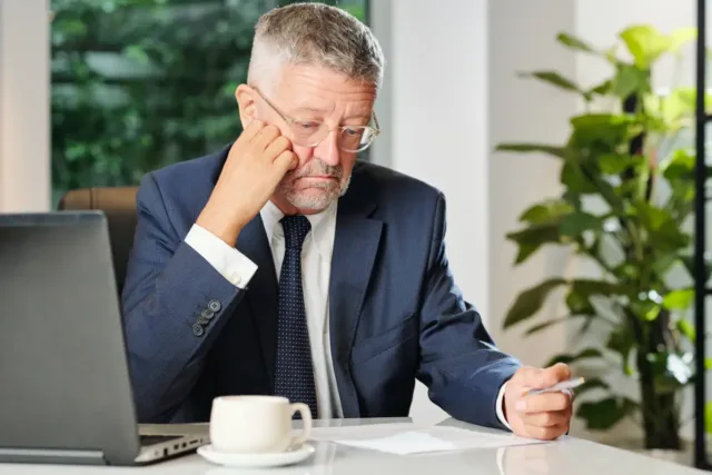 Mężczyzna w średnim wieku trzyma w ręce dokument, który czyta. Ma smutną minę, co może wskazywać na negatywną treść dokumentu.