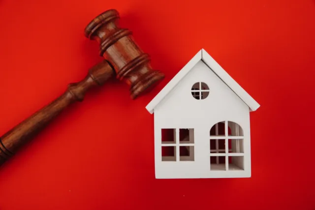 Młotek sędziowski skierowany w kierunku drewnianego domku. Może to symbolizować licytację komorniczą nieruchomości.